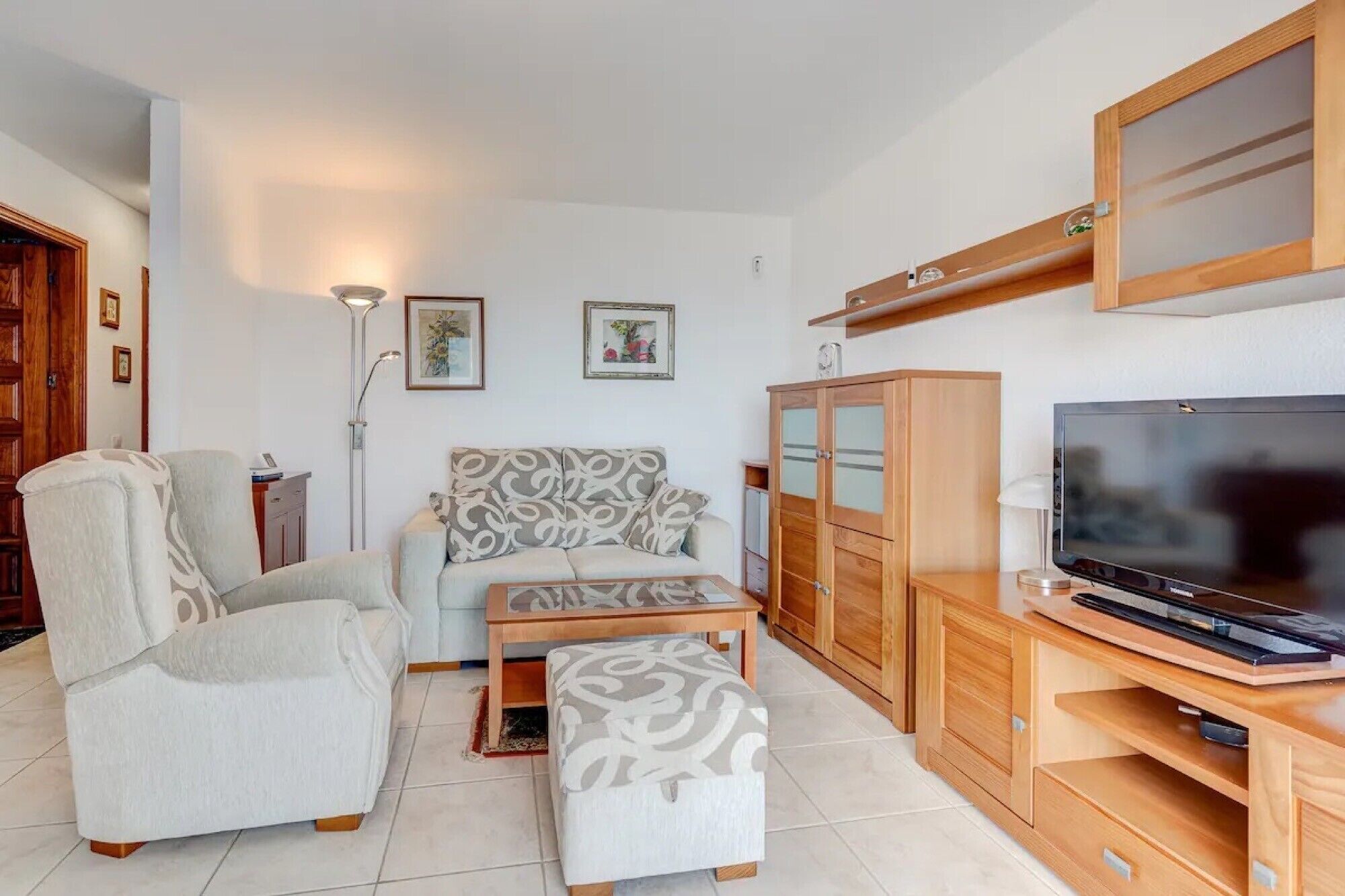 Ein Wohnzimmer mit einer Couch, einem Sessel, einem Hocker, einem Fernseher, einer Holzkommode und gerahmten Bildern an der Wand.