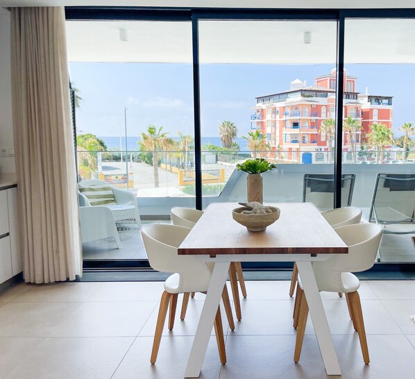 A dining area with a table and chairs, a vase with a plant, and a view of a building and palm trees through large windows.