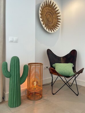 A room with a cactus, a woven lamp, a chair with a green cushion, and a wall-mounted decorative piece.
