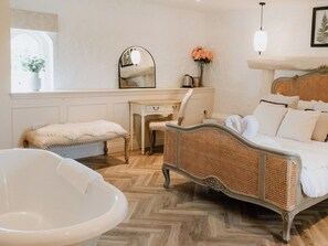 A bedroom with a large bed, a desk, a chair, a white bathtub, and a mirror.