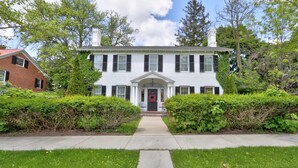 A two-story white house with black shutters, a front porch, and a red wreath on the door. - Historic Home: Game Room & Fire Pit, Near Downtown (Geneva)