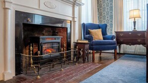 A room with a fireplace, a blue armchair, a wooden side table, and a lamp. - Historic Home: Game Room & Fire Pit, Near Downtown (Geneva)