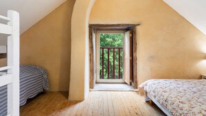 Une chambre avec un plafond en pente, une fenêtre donnant sur la verdure, un lit recouvert d’une couvre-lit à motifs floraux et un plancher en bois.