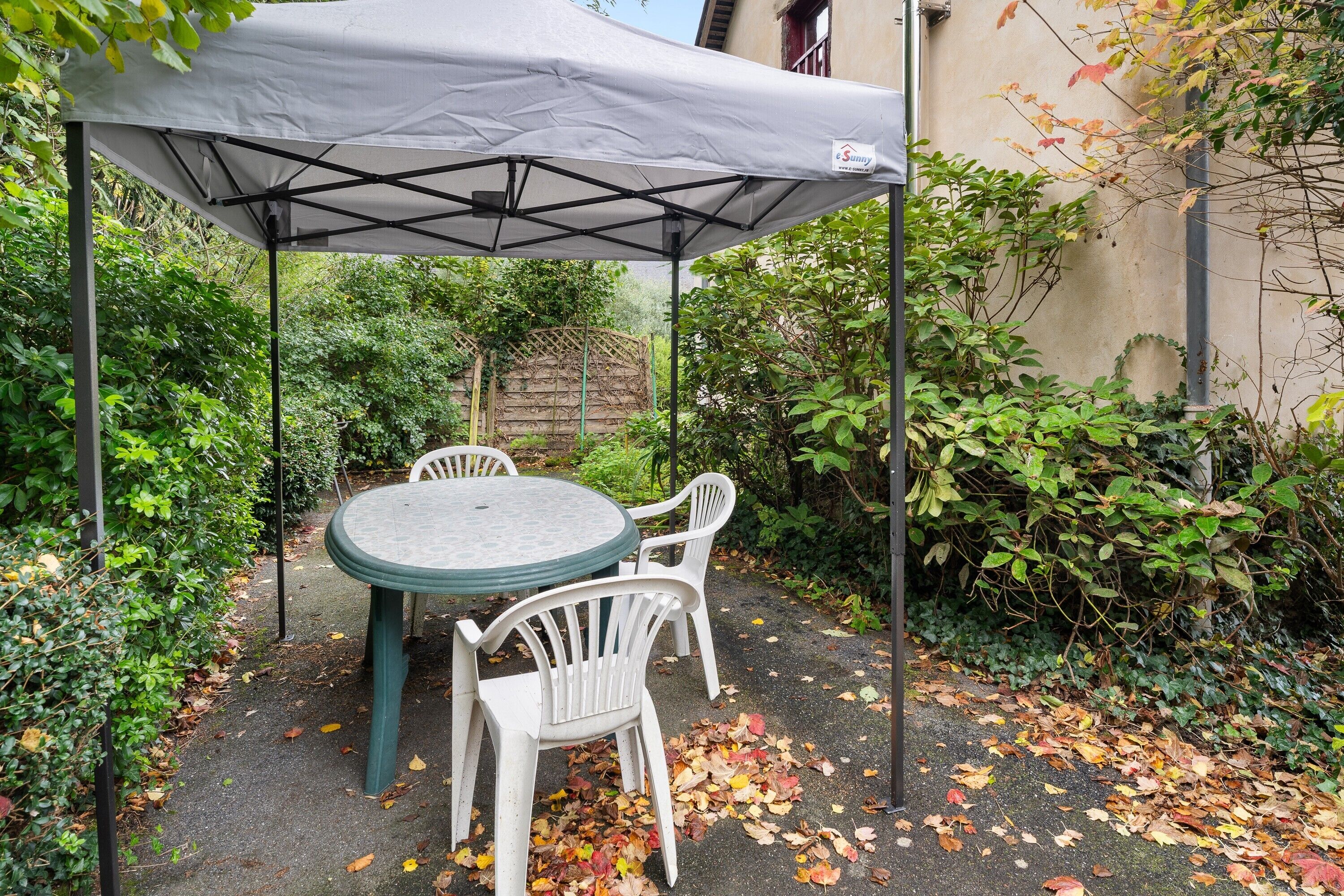 A patio with a table and chairs under a white canopy, surrounded by greenery.