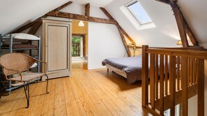Une chambre à coucher aménagée dans un grenier, avec un plafond en pente, des poutres en bois, un lit superposé, un lit simple, une chaise en osier et une armoire.