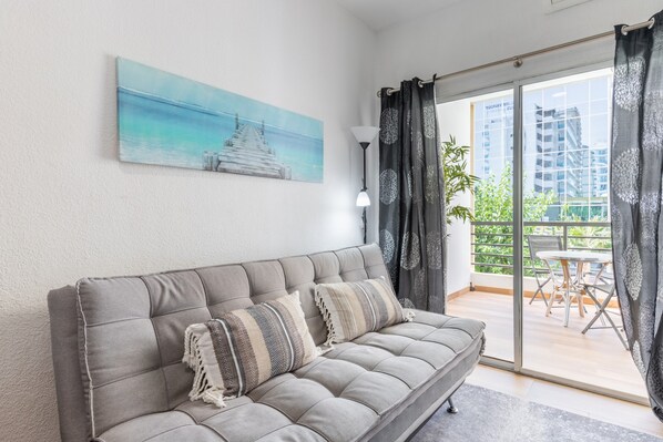 A modern living room with a grey sofa, a painting of a pier, and a view of a balcony with outdoor furniture.