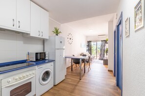 Een kleine keuken met een magnetron, oven en wasmachine. Een eethoek met een tafel en stoelen is aanwezig.
