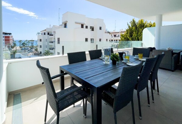 A balcony with a wooden table and chairs, a potted plant, and a clear view of buildings and the sea.