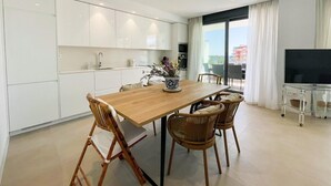 Une cuisine moderne avec des meubles blancs, une table à manger en bois, des chaises en osier et un téléviseur posé sur un meuble en verre.