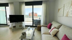 Un salon comprenant un canapé, une table basse, un téléviseur posé sur un meuble et une vue sur les immeubles à travers de grandes fenêtres.