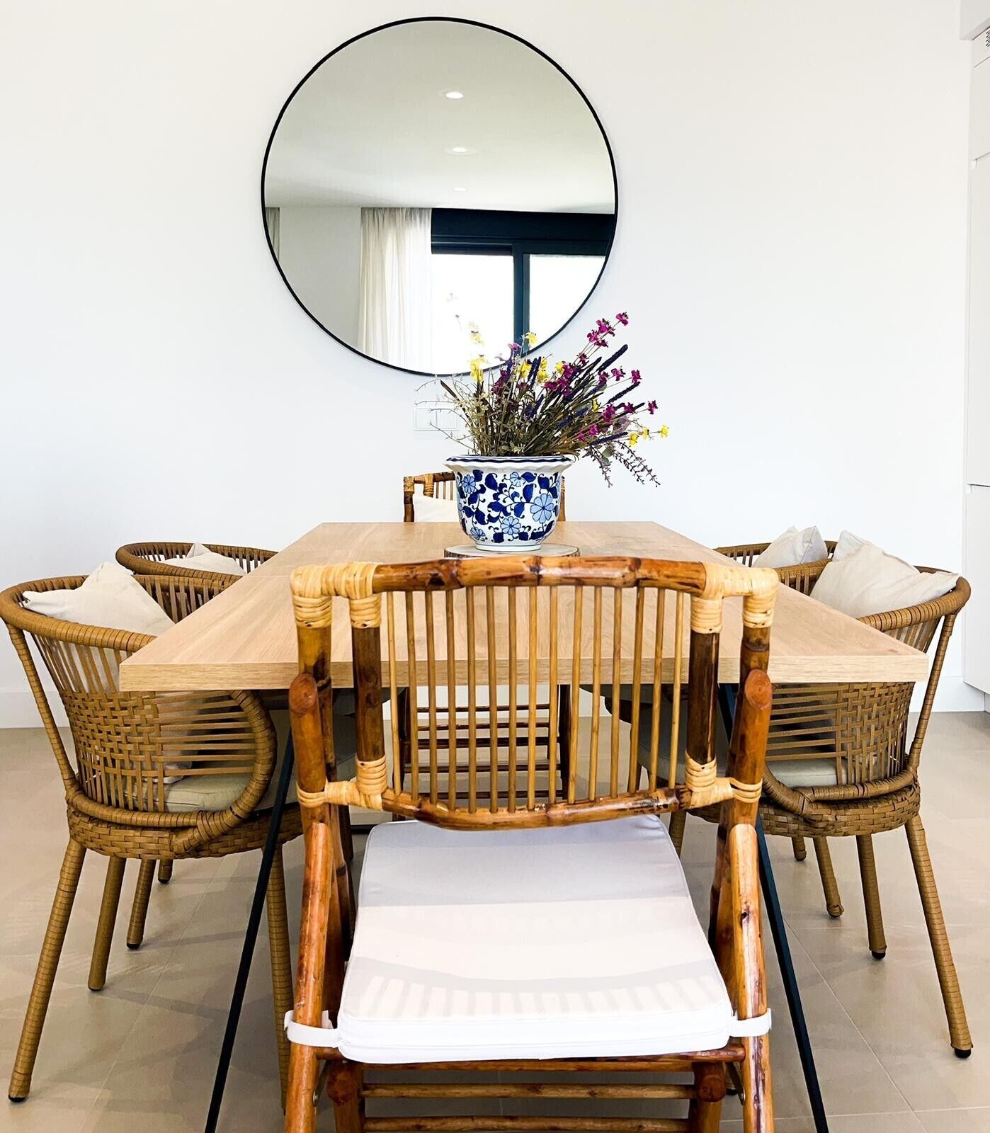 Uma área de refeições com uma mesa redonda, cadeiras de vime, um vaso com flores e um espelho grande na parede.