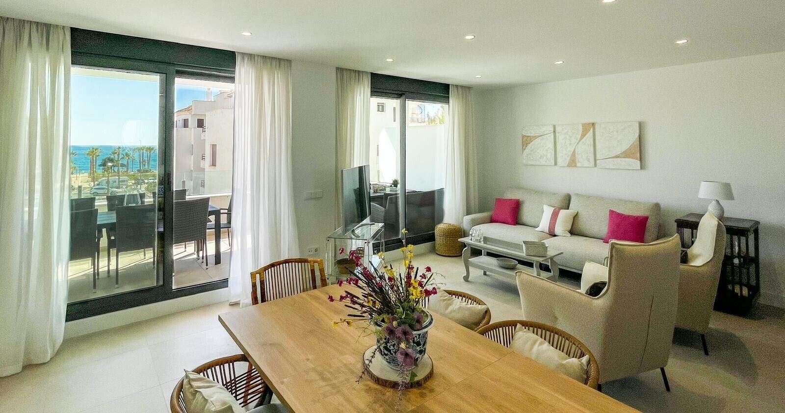 A modern living room with a dining table, sofa, and chairs, large windows with a view, and a flat-screen TV.