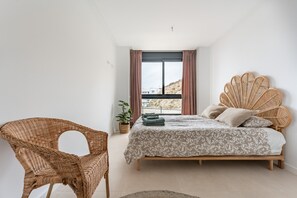 Een slaapkamer met een bed, een rieten stoel, een raam met gordijnen en een potplant.