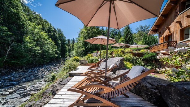 Une terrasse en bois, agrémentée de chaises longues et de parasols, entourée d’une zone boisée et d’un bâtiment doté d’un balcon.