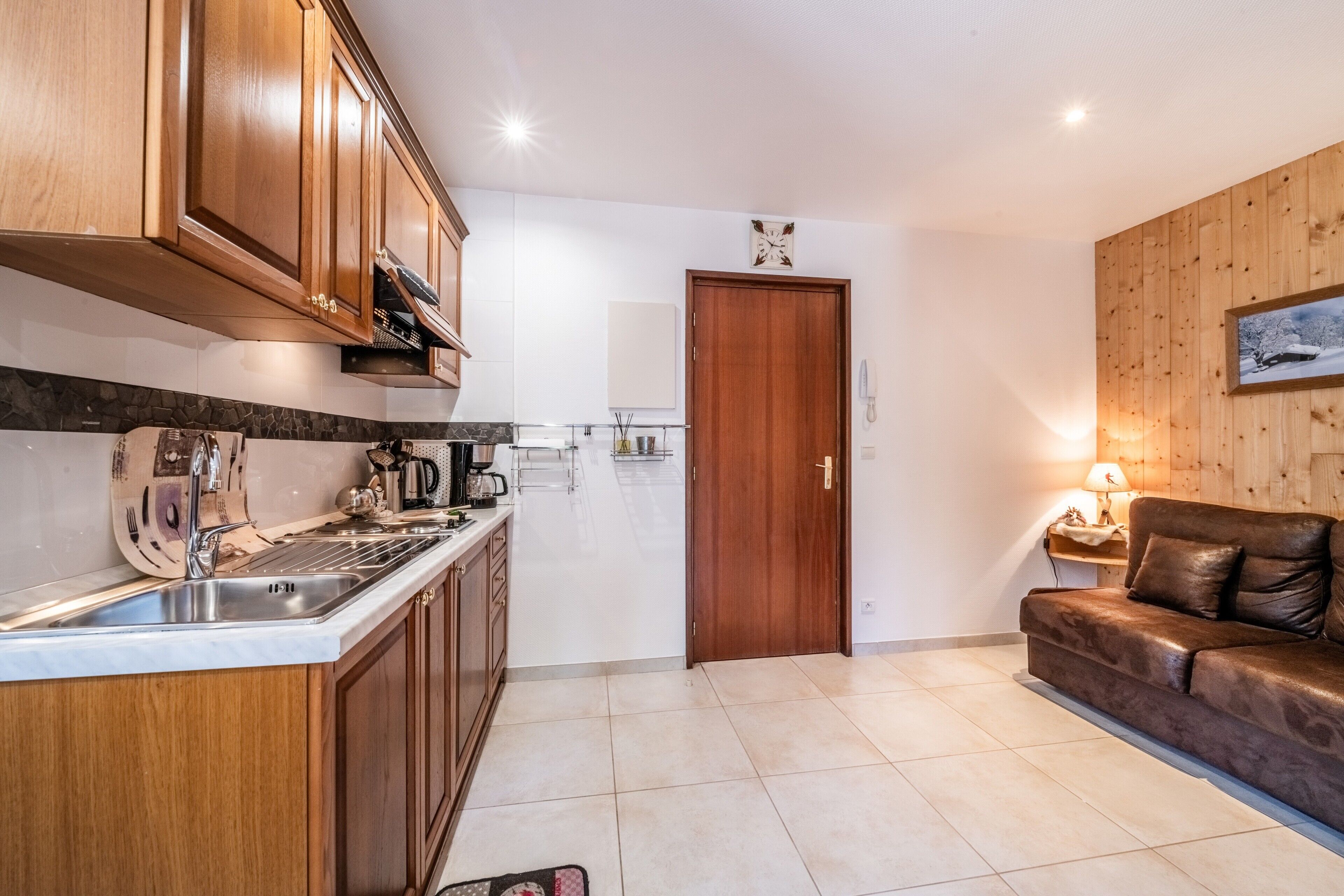 Een keuken met houten kastjes, een gootsteen en een aanrecht. In de woonkamer staat een houten deur en een bruine leren bank.