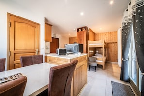 A compact kitchen with wooden cabinets, a central island with a toaster, a bed with a bunk, and a window with curtains.