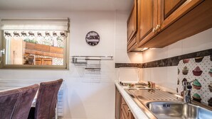 A kitchen with wooden cabinets, a stainless steel sink, a clock, and a window with a view of outdoor seating. - Studio 'Au Rêve Savoyard 1' with Shared Terrace, for 2 People (Morillon)