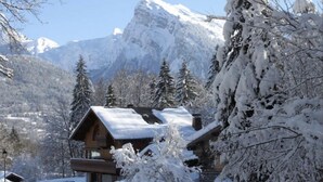 Una cabaña de madera con techo cubierto de nieve, rodeada de árboles cargados de nieve y montañas.