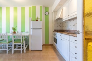 Una cocina con gabinetes blancos, un refrigerador blanco, un reloj en la pared y un comedor con mesa y sillas.