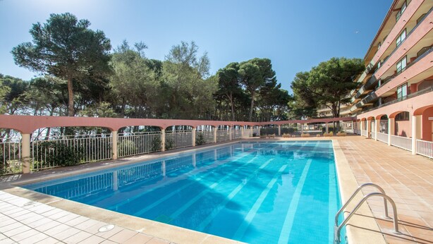 A swimming pool with a tiled deck, surrounded by trees and a building with balconies. - Apartment 'Salines' with Shared Pool, Balcony and Air Conditioning (Estartit)