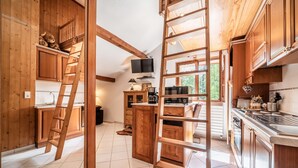 A kitchen with wooden cabinets, a central island, a TV mounted above the island, and a ladder leading to a lofted area. - Studio 'Au Rêve Savoyard 7' with Mountain View, for 4 People (Morillon)