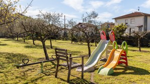 Un terrain de jeux avec une balançoire, un toboggan et une structure d’escalade, situé dans un espace verdoyant, avec des arbres et des maisons en arrière-plan.