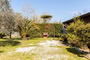 Un jardín con un sendero, diversas plantas y un edificio con tejado rojo.
