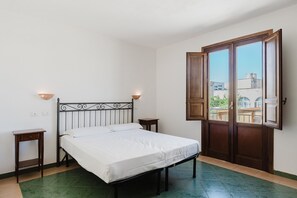 A bedroom with a bed, two bedside tables, a window with an open door, and a view of buildings outside.