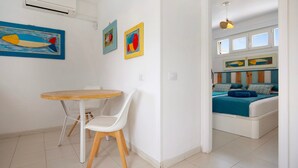 Ein modernes Wohnzimmer mit einem runden Holztisch und weißen Stühlen, einem Bett mit blauer Bettwäsche sowie Kunstwerken an den Wänden.