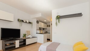 Une chambre moderne et bien éclairée, dotée d’un téléviseur à écran plat, d’un lit avec des oreillers jaunes, d’une petite kitchenette et d’un coin repas.