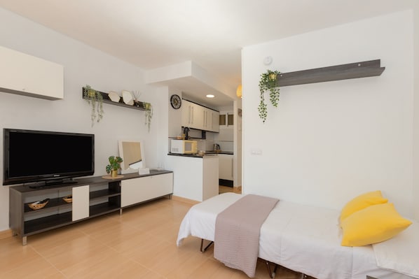 A modern, well-lit room with a flat-screen TV, a bed with yellow pillows, a small kitchenette, and a dining area.