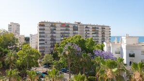 Un hôtel à plusieurs étages, doté de balcons, entouré de palmiers et de fleurs violettes, et offrant une vue sur une étendue d’eau.