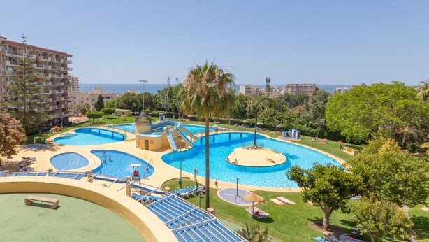 Une grande piscine extérieure divisée en plusieurs zones, entourée de verdure et offrant une vue sur la mer.