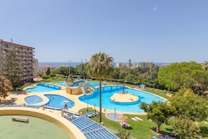 A large outdoor swimming pool with multiple sections, surrounded by greenery and a view of the sea.