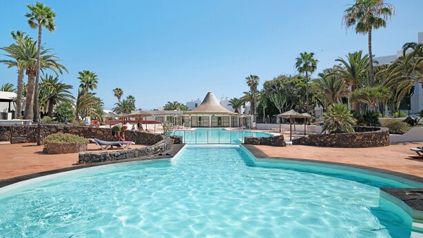 A swimming pool with lounge chairs, palm trees, and a building with a conical roof. - Vacation Apartment 'Al Sole Studios Playa Roca' with Sea View, Shared Pool and Wi-Fi (Costa Teguise)