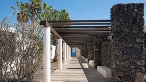A covered walkway with stone walls and palm trees. - Vacation Apartment 'Al Sole Studios Playa Roca' with Sea View, Shared Pool and Wi-Fi (Costa Teguise)