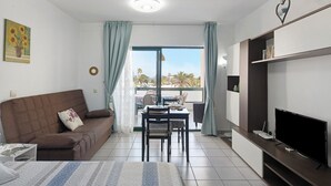 A modern living room with a sofa, a dining table, chairs, a flat-screen TV, and a balcony with a view. - Vacation Apartment 'Al Sole Studios Playa Roca' with Sea View, Shared Pool and Wi-Fi (Costa Teguise)