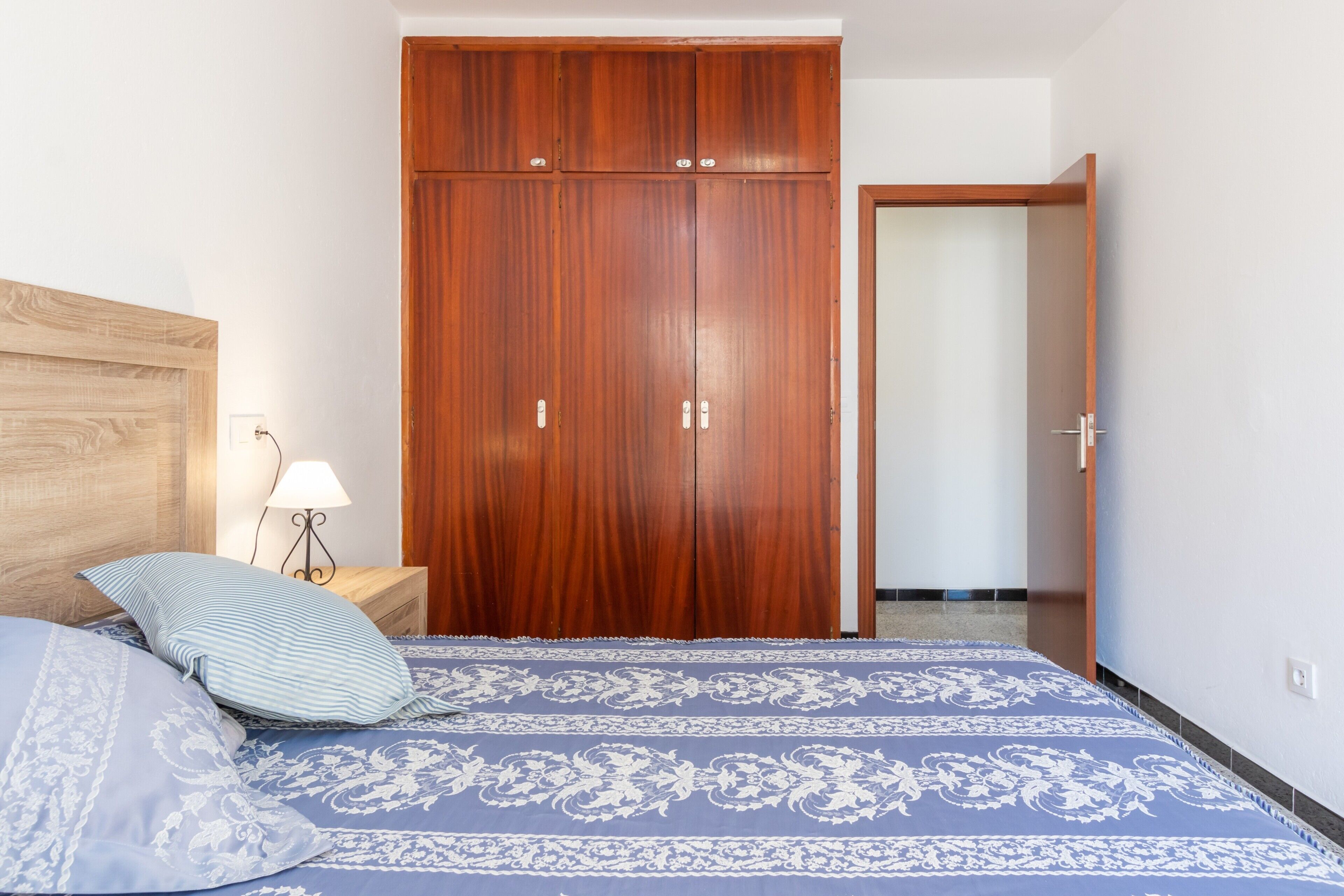 A bedroom with a wooden wardrobe, a bed with patterned bedding, a nightstand with a lamp, and a door leading to another room.