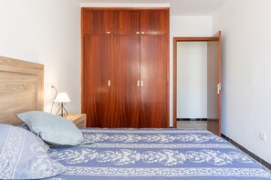 Ein Schlafzimmer mit Holzschrank, einem Bett mit gemusterten Betttextilien, einem Nachttisch mit Lampe und einer Tür zu einem anderen Raum.