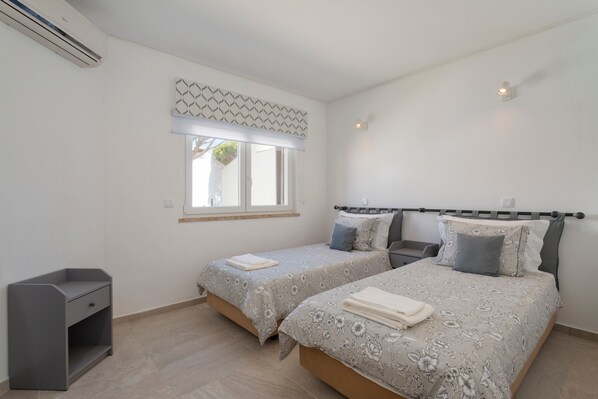 A bedroom with two beds, a window with a patterned valance, a bedside table, and a built-in air conditioning unit.