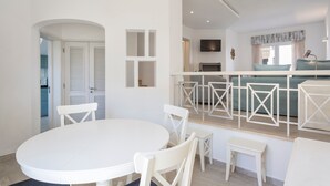 Une cuisine moderne avec des meubles blancs, un coin repas doté d’une table ronde et de chaises, et une vue sur le salon avec un canapé et un téléviseur.