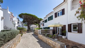 Un sentier pavé de pierres menant à un bâtiment blanc, avec un grand arbre, des murs en pierre et un parasol rayé.