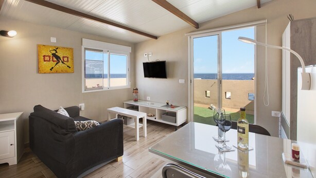 Ett modernt vardagsrum med en soffa, ett bord med glasskiva, en platt-TV och ett stort fönster med utsikt över en strand.