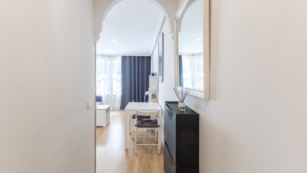 A narrow hallway leading to a dining area with a table and chairs, a black cabinet, and a large mirror on the wall.