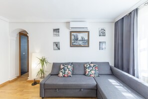 Een moderne woonkamer met een grijze bank, bloemenkussens, een potplant en ingelijste kunst aan de muur.