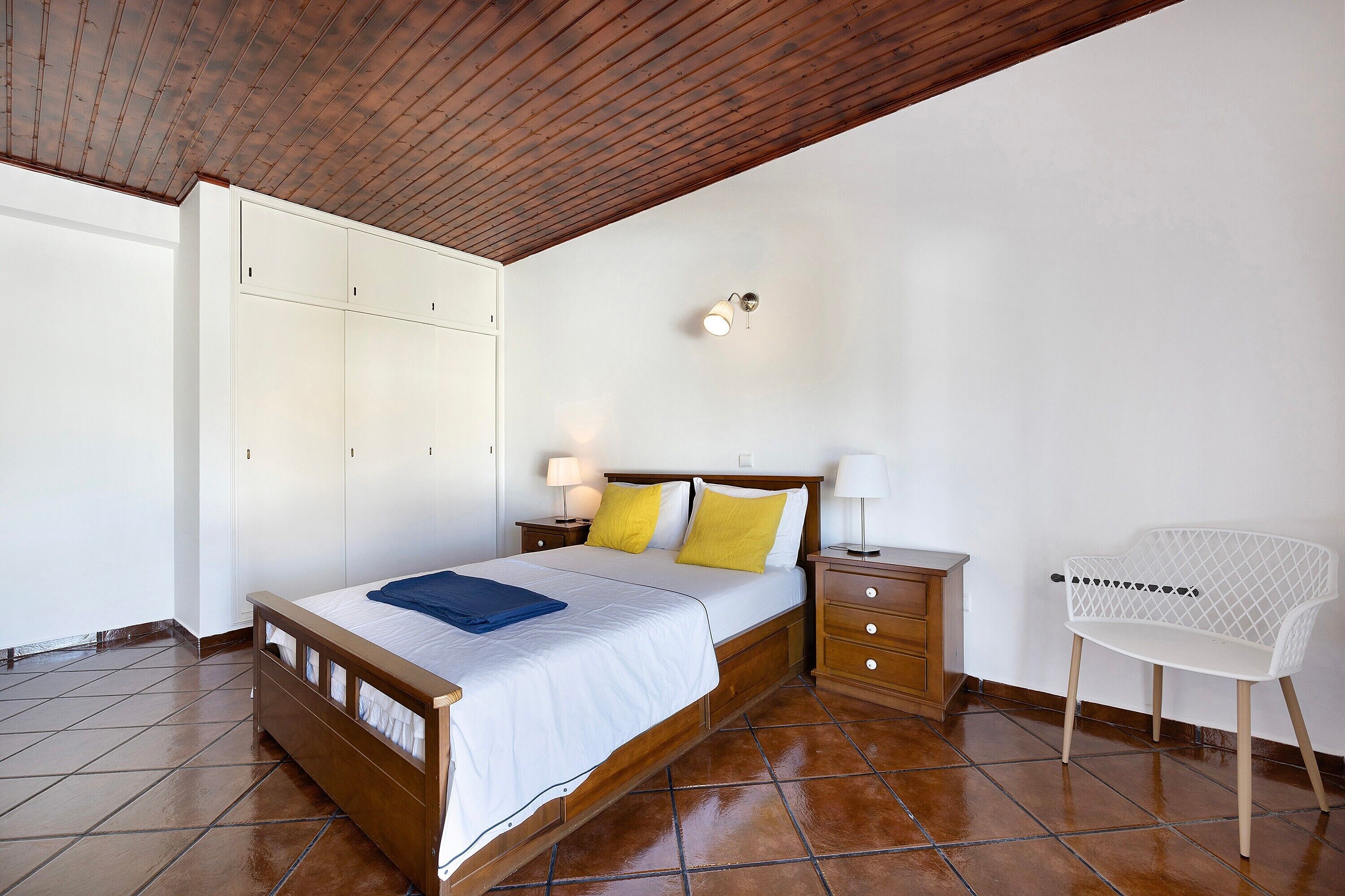 A bedroom with a wooden bed, a white wardrobe, a nightstand with a lamp, and a white chair.