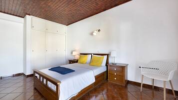 A bedroom with a wooden bed, a white wardrobe, a nightstand with a lamp, and a white chair.