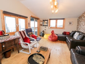 A cozy living room with a wooden floor, a leather sofa, a white armchair, and a red bean bag. There are windows with wooden frames, a stone wall, and a ceiling light fixture. - Sibrwd Y Mor (Criccieth)