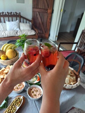 A person holding two glasses of a red beverage with ice and mint leaves, with a bowl of yellow fruit and a basket of bread on a table. - Historic Azorean Stable House | Private Patio & Farm Views (Lagoa, São Miguel Açores)