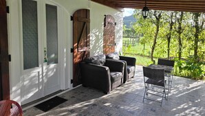 A covered patio with a view of a green landscape, a door with a glass panel, and a seating area with a chair and a sofa. - Historic Stable House | Private Patio & Farm Views (Lagoa, São Miguel Açores)